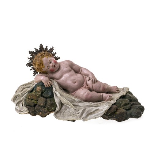 Schlafendes Christuskind. Terrakotta, polychrom gefasst. 18./19. Jahrhundert.Auf einer Decke liegendes Christuskind, das an einen Felsen gelehnt ist und schläft. Ca. 14 x 30 x 15,5 cm. Rest.