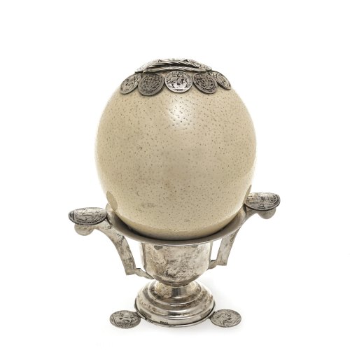 Straußenei als Sparbüchse. Als Halterung Silberschale mit Münzen. H. 22 cm.