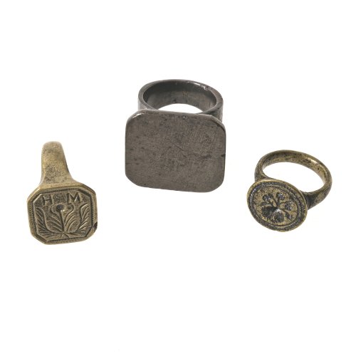 Drei Ringe. Messing. Einer mit Monogramm HM, einer mit Blütenmotiv, einer glatt, ungraviert. Alterungsspuren.